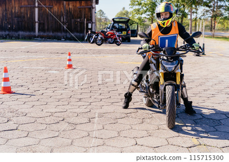 A person riding a motorcycle with the number 1, wearing a helmet, travels past a tree. The vehicles wheel and tire are visible in the image. Motorcycle driving school concept. A person riding a motorcycle with the number 1, wearing a helmet, travels past a tree. The vehicles wheel and tire are visible in the image. Motorcycle driving school concept. 115715300