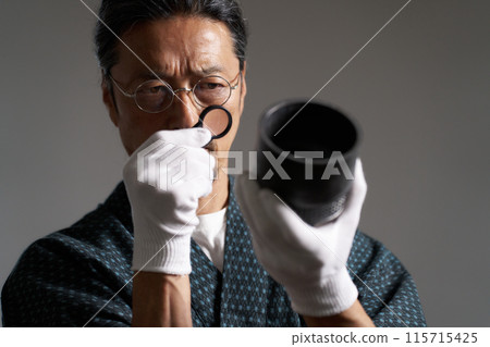一位身穿日本服裝的男性鑑賞家用放大鏡檢查陶瓷的圖像 一位身穿日本服裝的男性鑑賞家用放大鏡檢查陶瓷的圖像 115715425