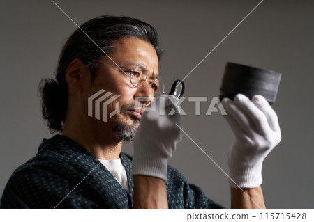 一位身穿日本服裝的男性鑑賞家用放大鏡檢查陶瓷的圖像 115715428