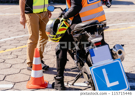 A man instructor in a yellow vest is standing next to a motorcycle with female student with a yellow helmet in hands. Letter L sign plate. Driving moto school driving. 115715446