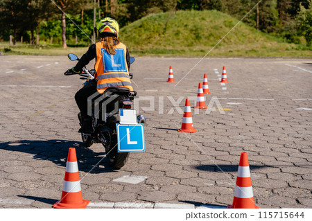 A woman is riding a motorcycle with a sign on the back that says L. The image features a street with asphalt, yellow lines, trees, and a vehicle for recreation and sports 115715644