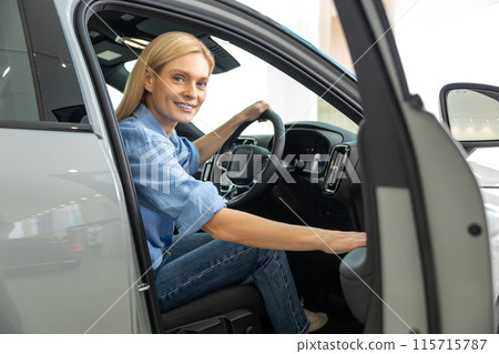 Blonde woman in a blue shirt sitting in a car and looking contented 115715787