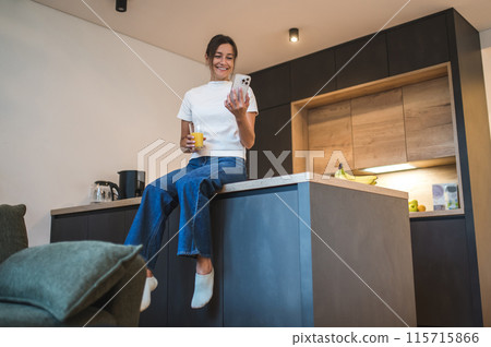 Cute young woman with a smartphone oh her kitchen at home 115715866
