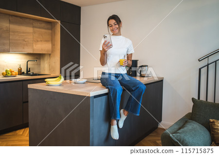 Cute young woman with a smartphone oh her kitchen at home Cute young woman with a smartphone oh her kitchen at home 115715875