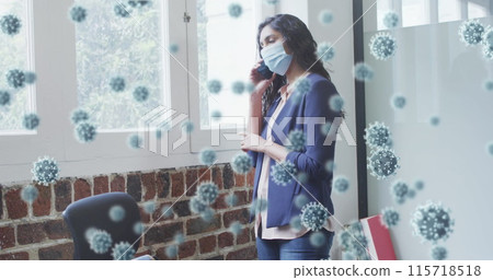 Image of covid 19 cells floating over man wearing face mask, using smartphone in office 115718518
