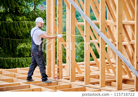 Carpenter constructing wooden-framed dwelling. Man measures distances with measuring tape while wearing work attire and headgear. The idea behind contemporary, eco-friendly building practices. Carpenter constructing wooden-framed dwelling. Man measures distances with measuring tape while wearing work attire and headgear. The idea behind contemporary, eco-friendly building practices. 115718722