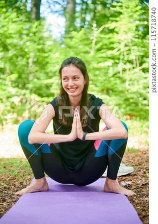Happy woman practicing yoga and meditating outdoors in the forest in a serene, natural setting. Smiling female performing yoga pose on mat. 115718740
