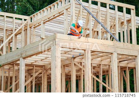 Carpenter constructing two-story wooden frame house. Bearded man hammering nails into structure while wearing protective helmet and construction vest. Concept of modern ecological construction. Carpenter constructing two-story wooden frame house. Bearded man hammering nails into structure while wearing protective helmet and construction vest. Concept of modern ecological construction. 115718745