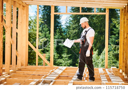 Supervisor overseeing construction of wooden two-story house located near woods. Bearded man dressed in jumpsuit and protective helmet is studying the construction plan. Supervisor overseeing construction of wooden two-story house located near woods. Bearded man dressed in jumpsuit and protective helmet is studying the construction plan. 115718750