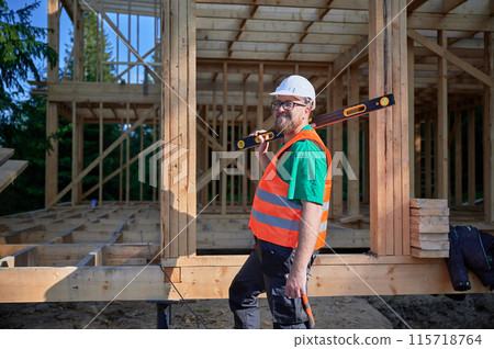 Carpenter building wooden frame two-story house. Bearded man in glasses holding a level, wearing protective helmet, overalls and orange vest. Concept of modern ecological construction. 115718764