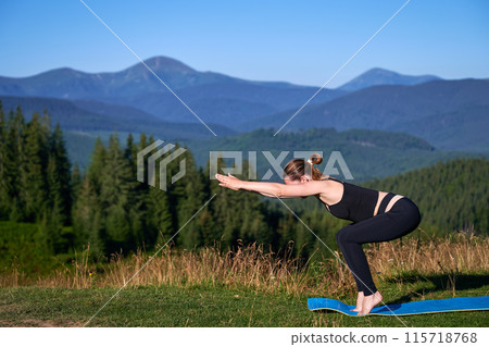 Woman practicing yoga outdoors in the mountains in a serene, natural setting. Female performing yoga pose on blue mat, with backdrop of beautiful mountain landscape at sunrise or sunset. 115718768