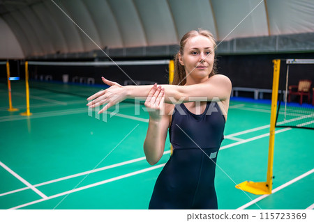 Sports young woman with racket and shuttlecock is exercising, doing warm-up before playing in badminton on inside court 115723369