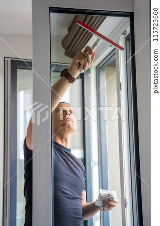 Young man cleans the windows in the house. He splashes detergent, rubs and removes water at the glass Young man cleans the windows in the house. He splashes detergent, rubs and removes water at the glass 115723660