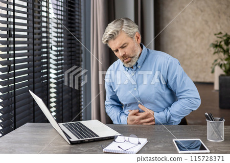 Businessman experiencing stomach pain while working on a laptop at a modern office desk. Stress, health issues, and discomfort. 115728371