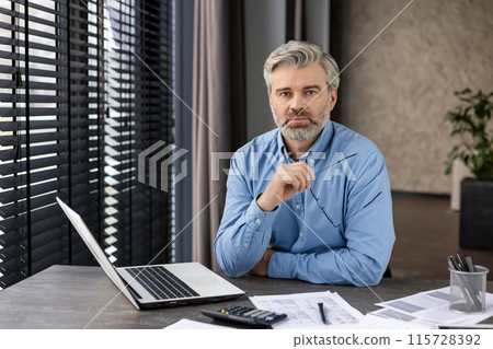 Confident businessman sitting at desk, holding glasses, working on laptop, and analyzing documents in modern office Confident businessman sitting at desk, holding glasses, working on laptop, and analyzing documents in modern office 115728392