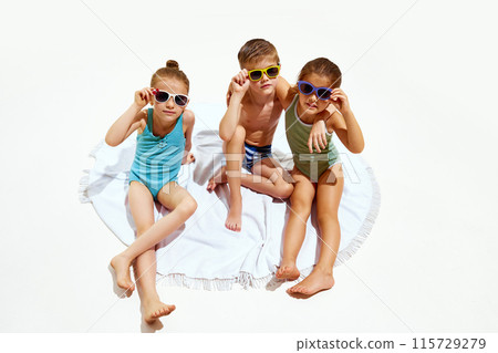 Cute little children, girls and boy in swimsuits relaxing on beach towel against white studio background. 115729279