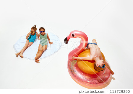 Cute little girls in swimsuits relaxing on beach towel, and boy lying atop orange exercise ball on flamingo float against white studio background. Cute little girls in swimsuits relaxing on beach towel, and boy lying atop orange exercise ball on flamingo float against white studio background. 115729290