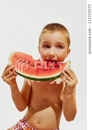 Portrait of cute little boy sitting in swimwear and eating huge slice of watermelon against white studio background. 115729337
