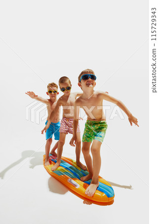 Three boys in swim trunks and sunglasses balancing, as surfing on inflatable board against white studio background. Three boys in swim trunks and sunglasses balancing, as surfing on inflatable board against white studio background. 115729343