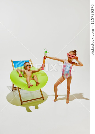 Kids have fun on sunny day. Boy relaxes in beach chair with green inflatable ring, while his friend, girl holds toy water blaster. 115729376
