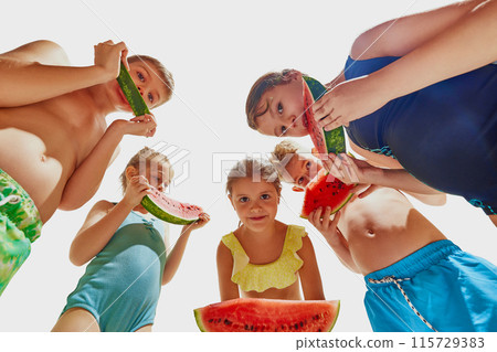 View from below of cute little children, boys and girls in swimsuits eating red, sweet watermelon against white studio background. 115729383
