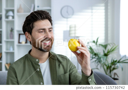 Close-up photo of a smiling young man in a green shirt at home, holding and looking at an apple. Close-up photo of a smiling young man in a green shirt at home, holding and looking at an apple. 115730742