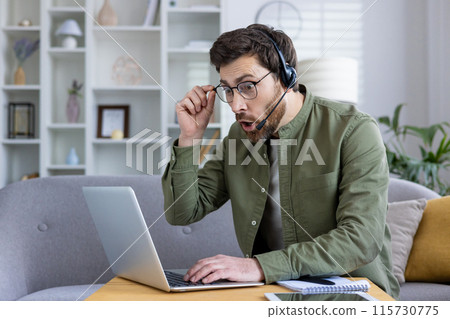 Surprised man in glasses wearing a headset while using a laptop in a home office environment. Concept of remote work, communication, and unexpected news 115730775