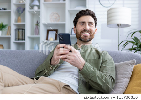Man smiling while using a smartphone, relaxing on a couch in a modern, stylish living room. He appears content and comfortable. Man smiling while using a smartphone, relaxing on a couch in a modern, stylish living room. He appears content and comfortable. 115730810