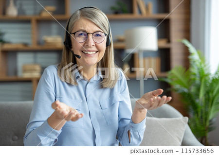Senior woman wearing a headset during a video call, smiling and talking happily, sitting in a cozy living room. 115733656