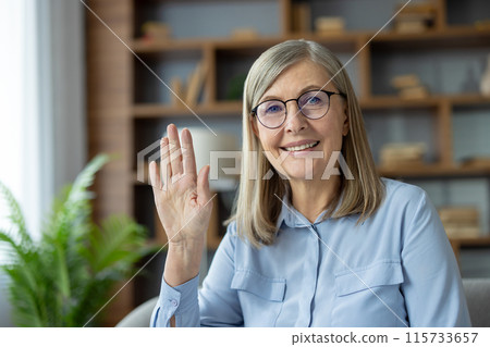 Senior woman with glasses smiling and waving while engaging in a video call from home, representing communication and connection. Senior woman with glasses smiling and waving while engaging in a video call from home, representing communication and connection. 115733657