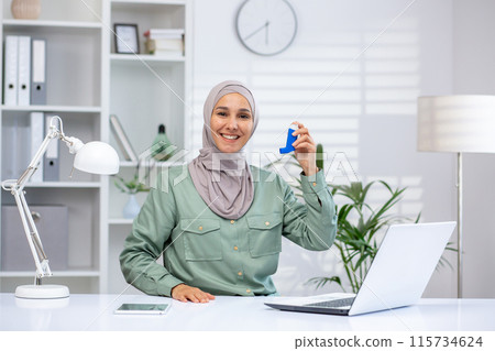 Smiling woman with hijab holding an inhaler at her desk in a modern office, promoting health and wellness. Smiling woman with hijab holding an inhaler at her desk in a modern office, promoting health and wellness. 115734624