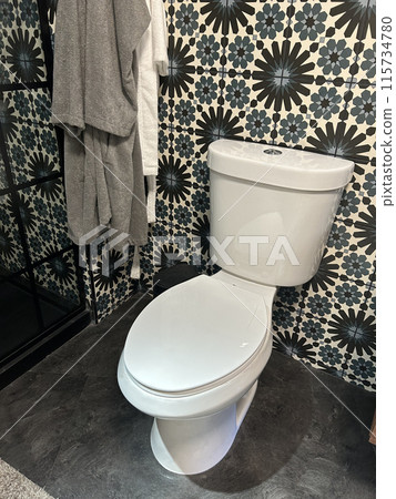 A modern bathroom features a white toilet, patterned tile wall, and hanging towels 115734780