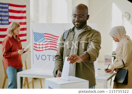Confident young Black male citizen placing ballot paper into ballot box at polling place on presidential election day 115735322