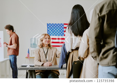 Mature Caucasian woman sitting at desk working at polling station on election day, giving ballot papers to voters Mature Caucasian woman sitting at desk working at polling station on election day, giving ballot papers to voters 115735398