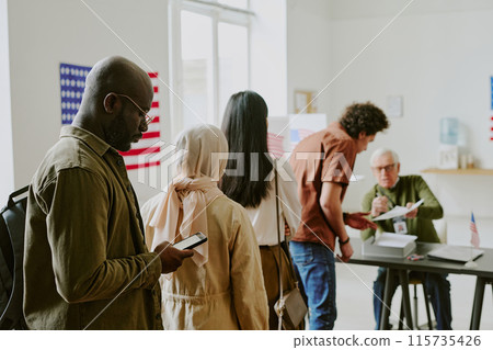 Young African American male citizen holding smartphone standing in line at voting station on election day, copy space Young African American male citizen holding smartphone standing in line at voting station on election day, copy space 115735426