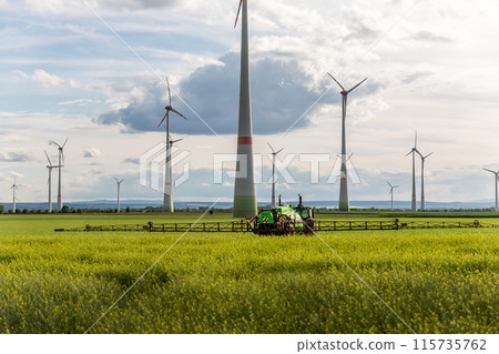 Scenic landscape view big modern tractor machine with sprayer equipment spraying fertilizer in rapeseed agricultural field against windfarm power windmill. Arable land cultivation sustainable energy Scenic landscape view big modern tractor machine with sprayer equipment spraying fertilizer in rapeseed agricultural field against windfarm power windmill. Arable land cultivation sustainable energy 115735762