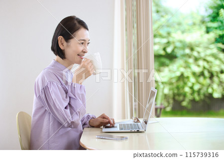 中年婦女在客廳喝咖啡時看著電腦 中年婦女在客廳喝咖啡時看著電腦 115739316