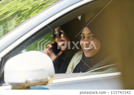 Two Muslim women wearing hijab converse on a smartphone while traveling together in a car through the  115741542