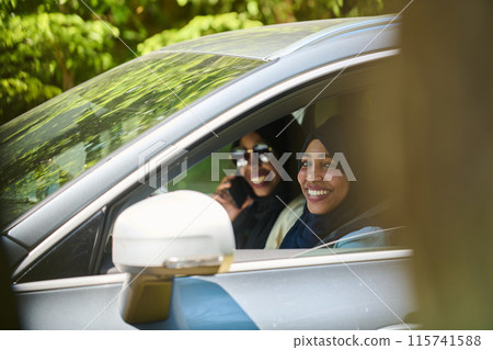 Two Muslim women wearing hijab converse on a smartphone while traveling together in a car through the  115741588