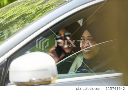 Two Muslim women wearing hijab converse on a smartphone while traveling together in a car through the  115741642