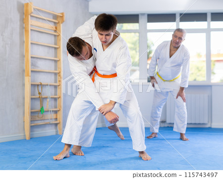 Two men practice grabbing and throwing sports mats during judo training under the guidance of an experienced mature coach 115743497