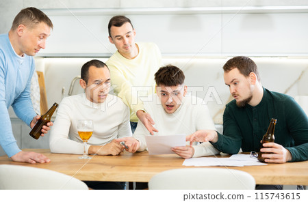 Male friends read a financial or legal document, drink beer and try to solve financial problems by consulting each other while sitting at table Male friends read a financial or legal document, drink beer and try to solve financial problems by consulting each other while sitting at table 115743615