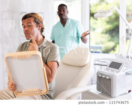 Man dissatisfied with results of rejuvenating at cosmetologist and meticulously examines reflection 115743675