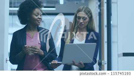 Two female colleagues are discussing something while walking and using laptops in an office setting 115745193