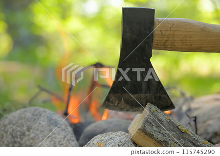 Axe lodged in a log of wood, positioned across from a campfire 115745290