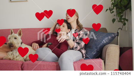 Image of hearts over caucasian female couple sitting on sofa with their pet dog 115748404