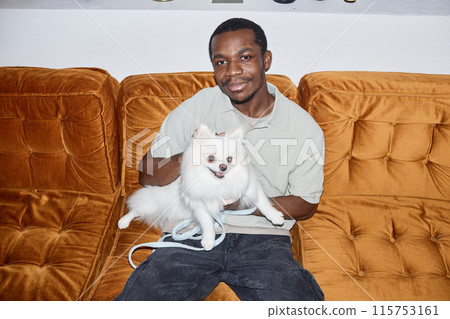 Portrait of smiling African American man holding white Spitz puppy sitting on velvet couch and looking at camera shot with camera flash copy space Portrait of smiling African American man holding white Spitz puppy sitting on velvet couch and looking at camera shot with camera flash copy space 115753161