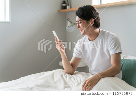 A young Asian man uses his smartphone to search and operate after waking up in bed at home or in a hotel. 115755975