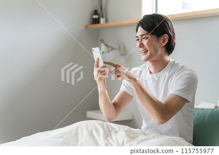 A young Asian man uses his smartphone to search and operate after waking up in bed at home or in a hotel. A young Asian man uses his smartphone to search and operate after waking up in bed at home or in a hotel. 115755977