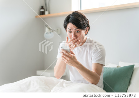 A young Asian man is surprised when he looks at his smartphone on the bed at home/hotel (surprised/late/shocked/dangerous) A young Asian man is surprised when he looks at his smartphone on the bed at home/hotel (surprised/late/shocked/dangerous) 115755978